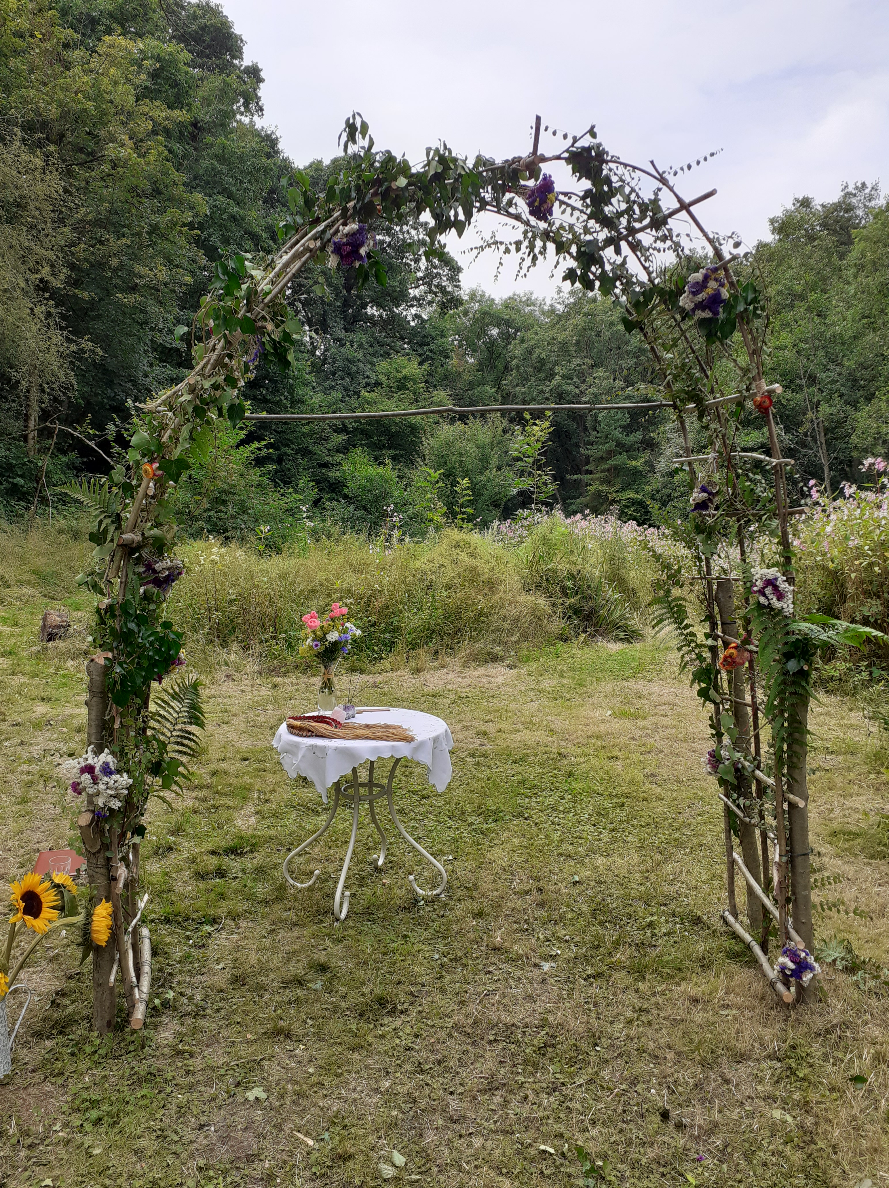 A DIY flower arch for an outdoor garden wedding. Beside it is a bucket of sunflowers and behind is an altar table with a Handfasting cord and vase of flowers. Taken by Keli Tomlin