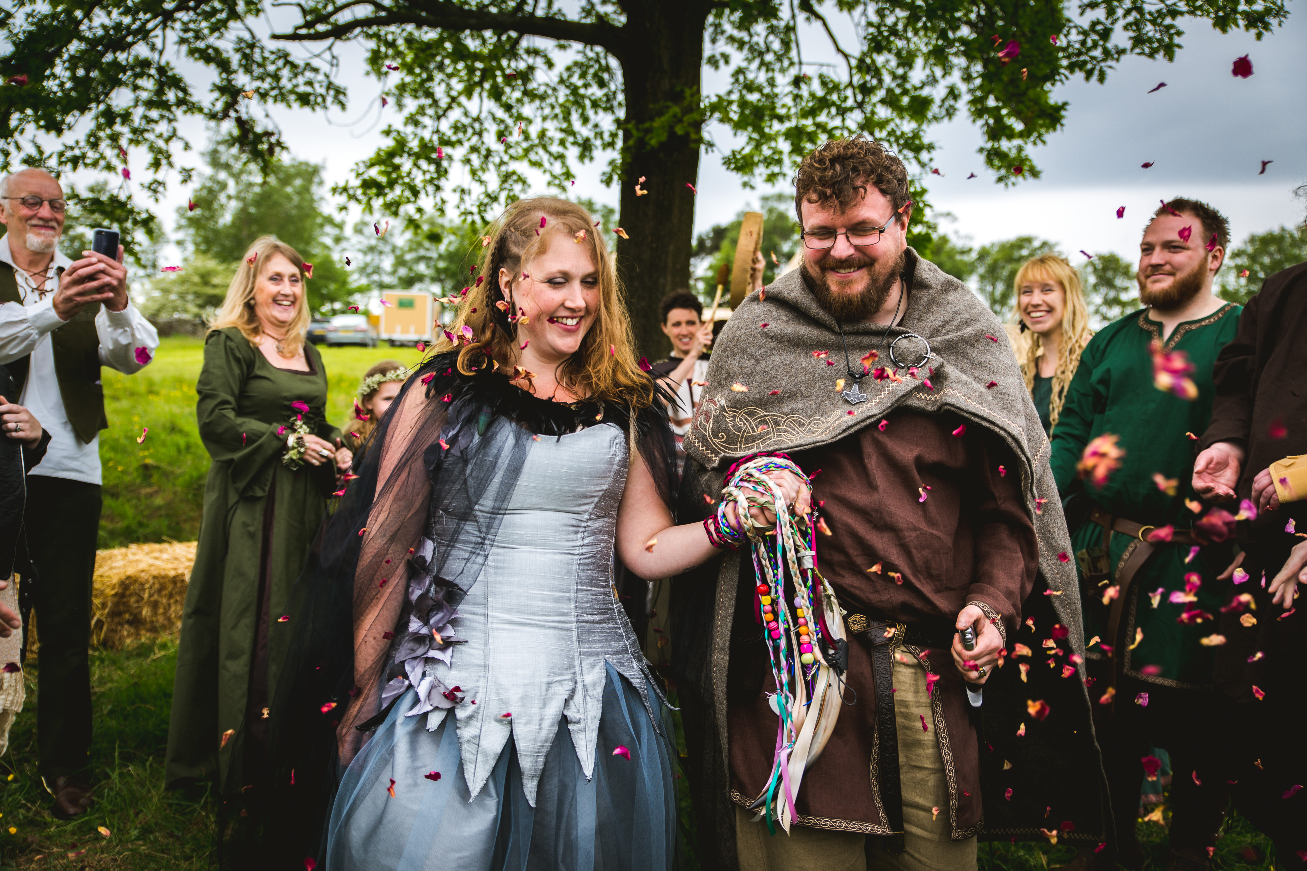 a viking bride and groom walk through their guests who throw confetti. theire hands are tied with handfasting cords and their Celebrant Keli Tomlin drums in the background to signal the end of their outdoor wedding ceremony