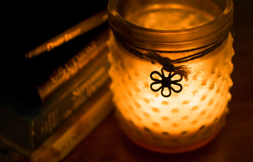 A teaclight candle lights up a glass holder which had dots on the frosted glass and a small daisy charm attached to sting around the jar rim. Beside the jar is a pile of books.